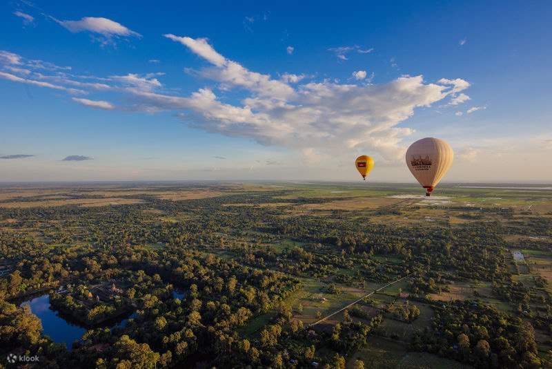 Aerial view of hot air balloons flying over lush green forests and rural landscapes during sunrise in Siem Reap.