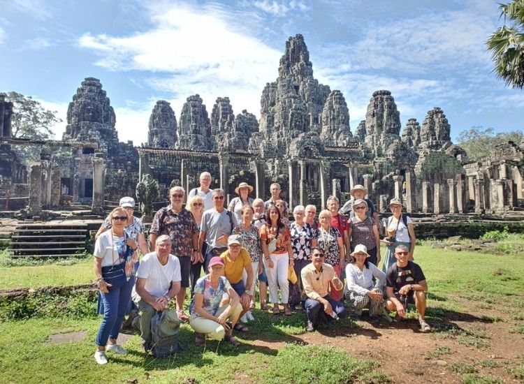 A large group of international tourists posing on the grassy lawn in front of the stone face towers of Bayon temple in Siem Reap.