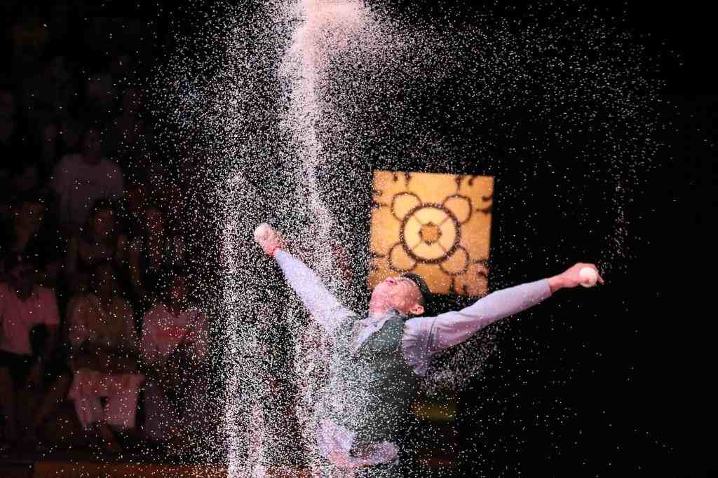 A Phare Circus performer standing on stage with arms open wide under a shower of white sand or powder during a theatrical show in Siem Reap.