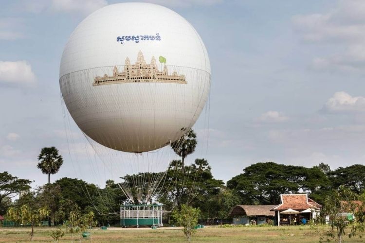 A large white helium balloon with an Angkor Wat illustration floating over trees and rural buildings in Siem Reap.