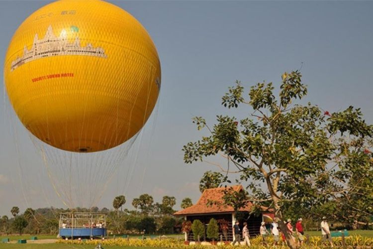 A large yellow tethered hot air balloon preparing for takeoff over trees and a traditional pavilion in Siem Reap.