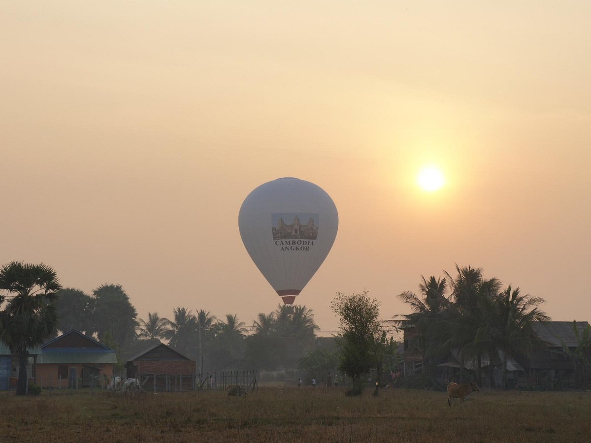 A passenger hot air balloon floating over a rural village and sugar palm trees during a hazy sunrise in Siem Reap.