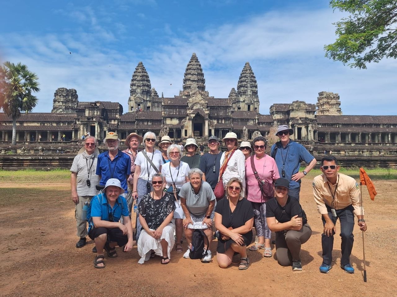 A group tour of international tourists posing together on the dirt path in front of the central stone towers of Angkor Wat temple in Siem Reap.