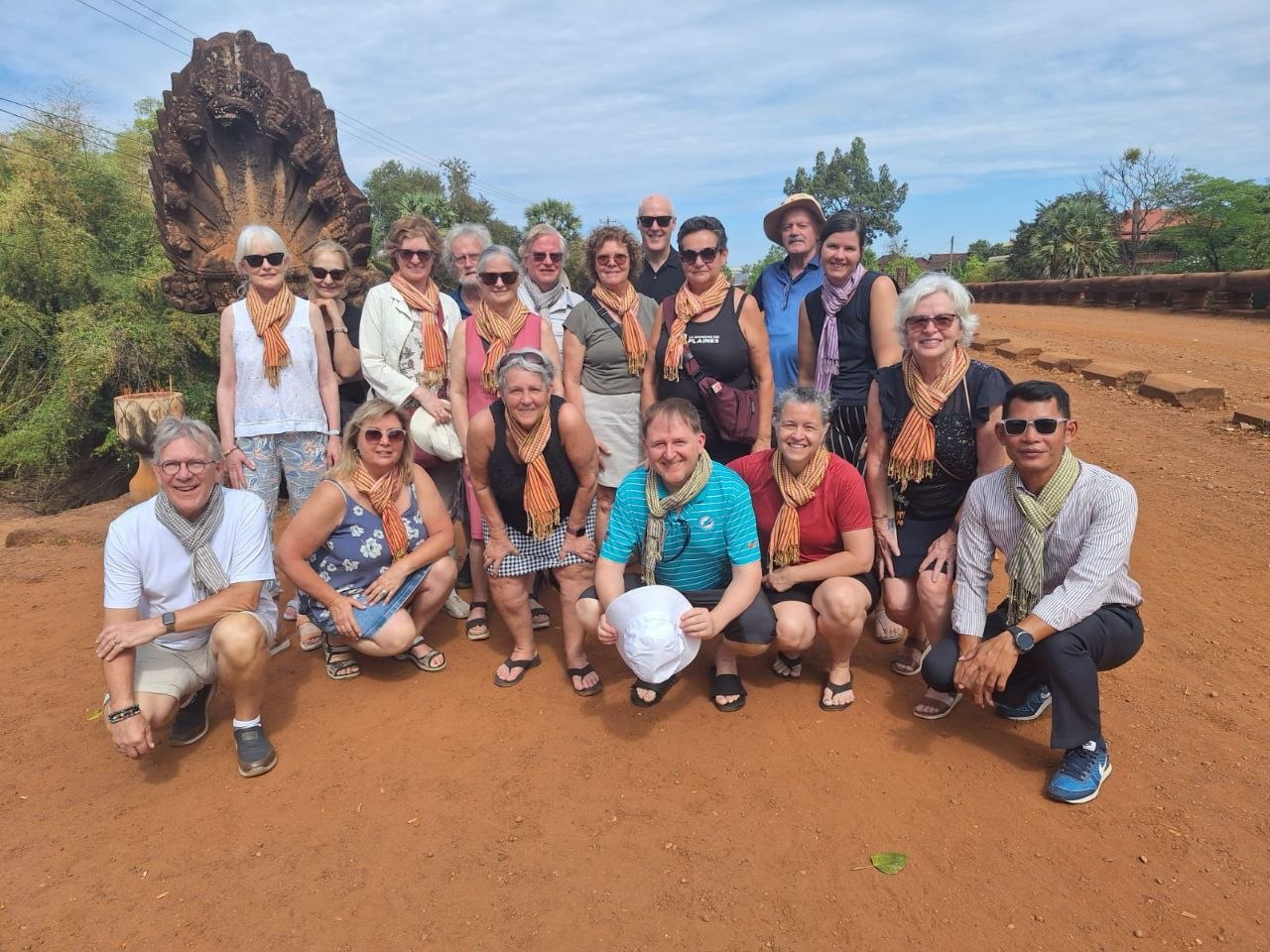 A large group of smiling senior tourists posing on red dirt grounds with their local guide during a temple tour in Siem Reap.