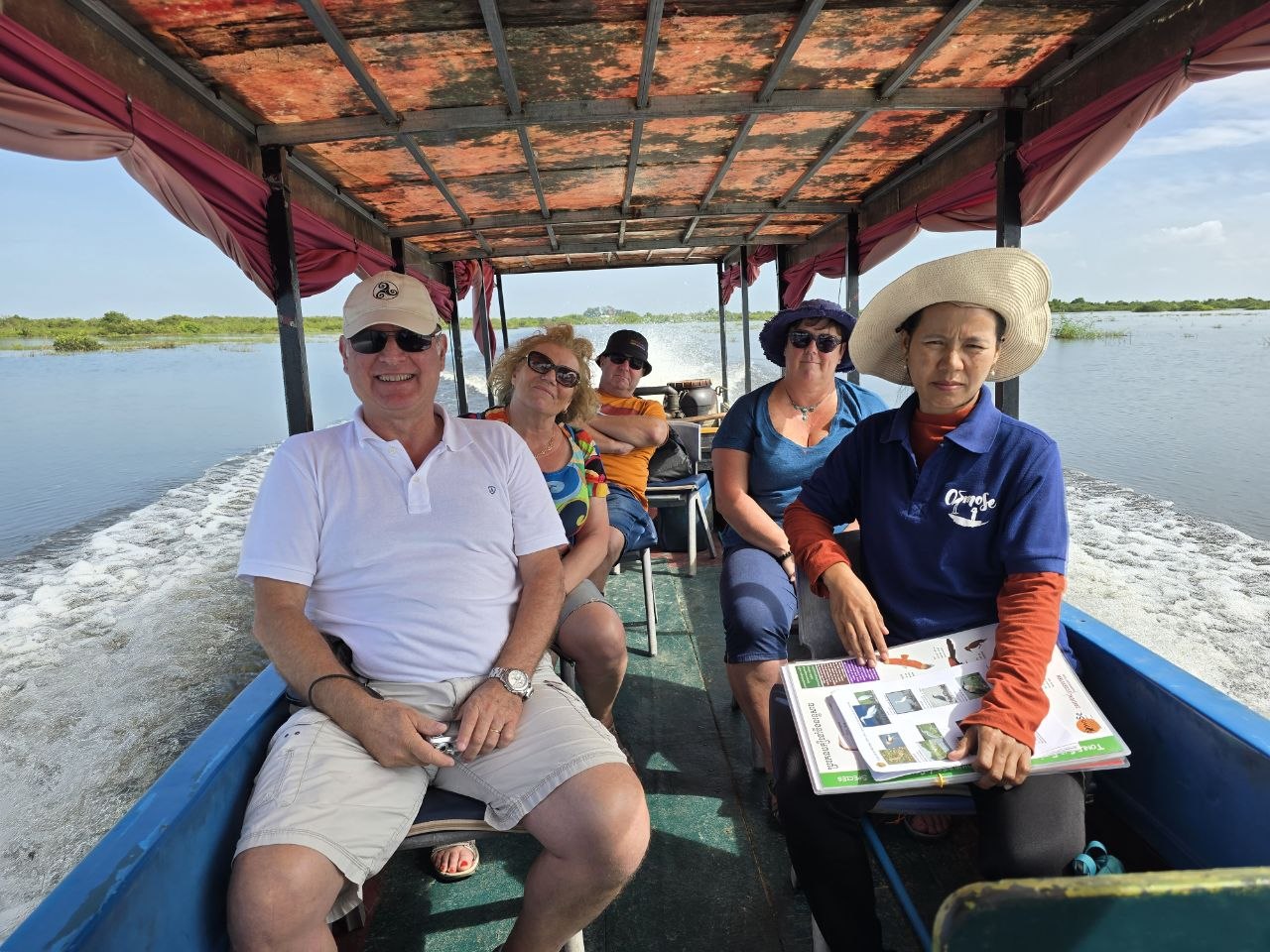 A group of tourists sitting on a covered wooden boat with their local female tour guide during a floating village excursion in Siem Reap.