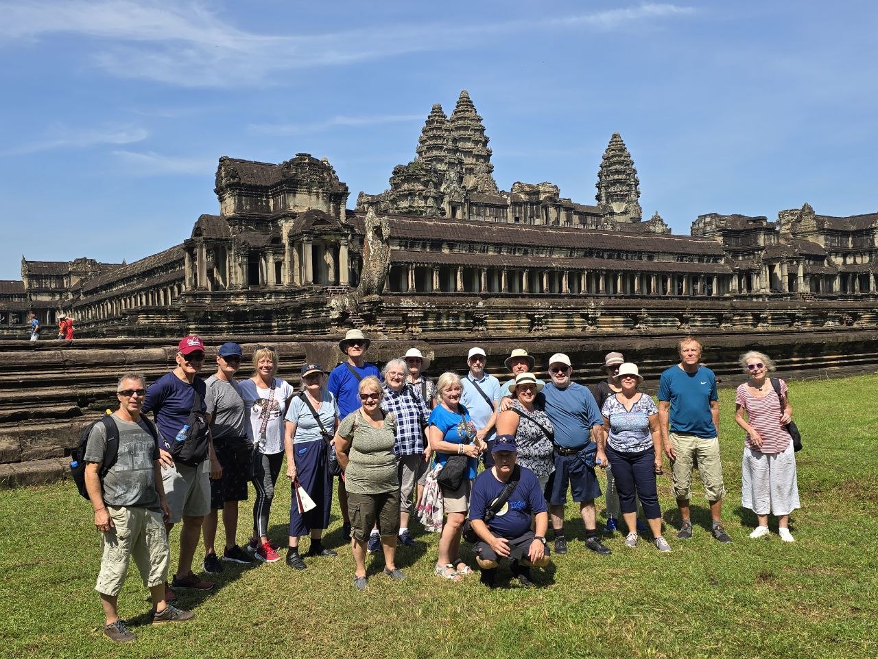 A large group tour of international senior tourists posing together on the green grass outside Angkor Wat temple in Siem Reap.