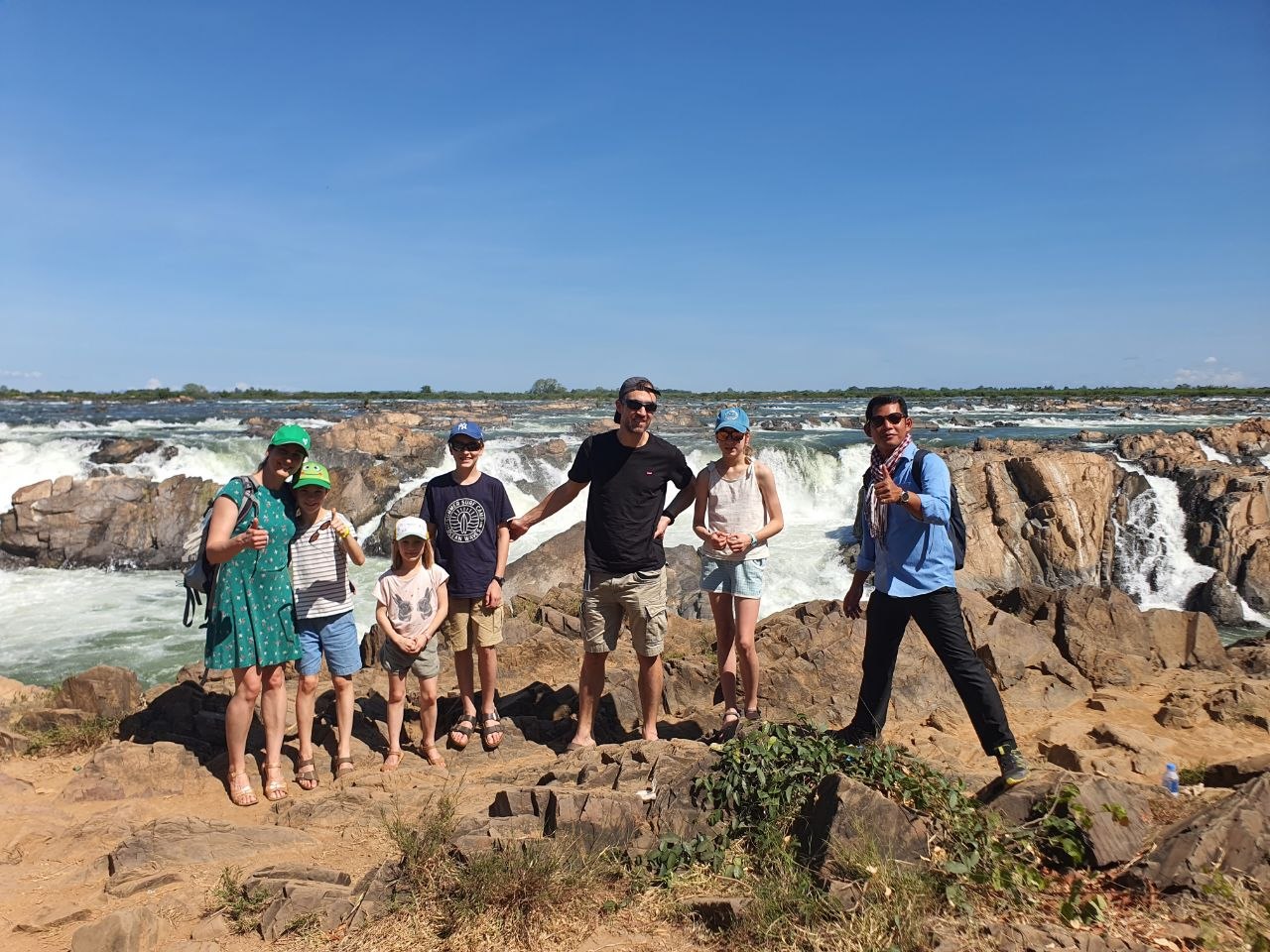 A family of tourists posing on a rocky overlook in front of a wide rushing waterfall with their local tour guide.