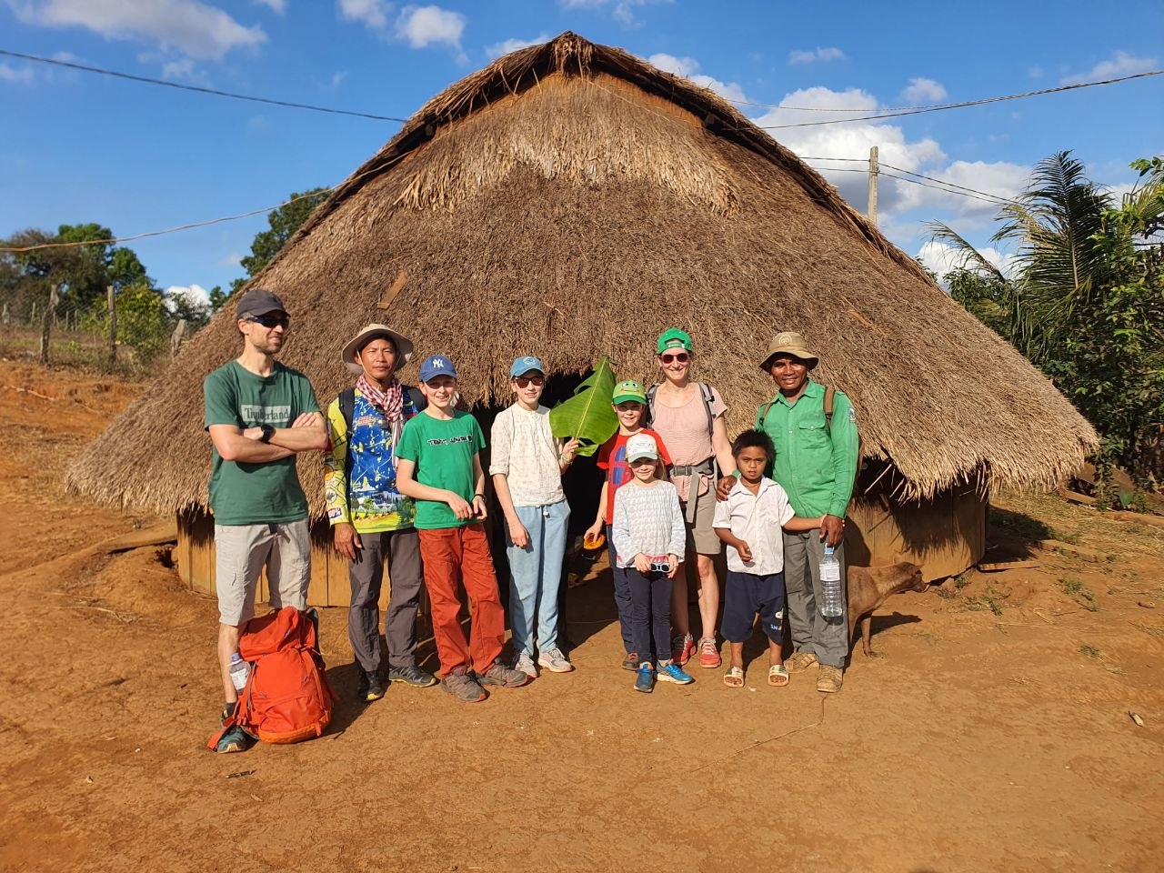 A family of tourists posing with a local family outside a traditional thatched-roof hut in a rural village near Siem Reap.