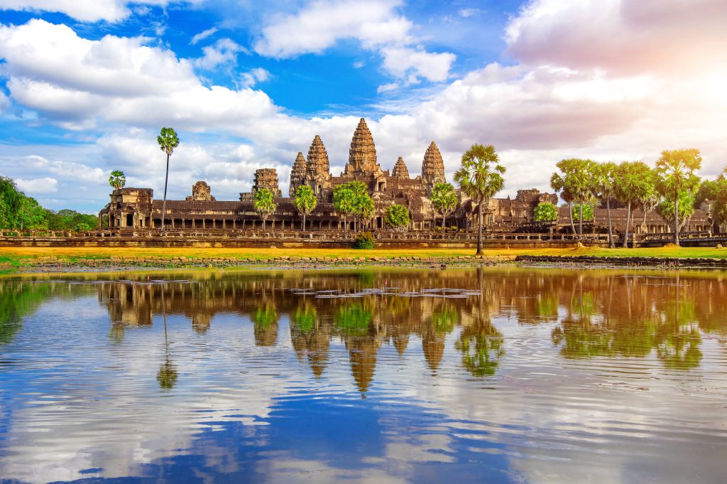 Wide panoramic view of Angkor Wat temple and its central stone towers perfectly reflected in the calm reflection pond under a bright sky in Siem Reap.