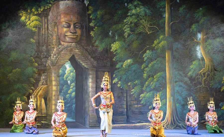 A group of Cambodian Apsara dancers performing on a stage set designed like a stone temple gateway in the jungle.