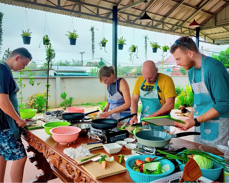 Tourists wearing aprons chop fresh vegetables at outdoor cooking stations during a Cambodian cooking class in Siem Reap.