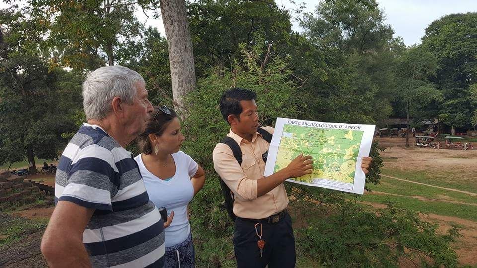 A local Cambodian tour guide holding an Angkor archaeological map while explaining the layout to two tourists outdoors.