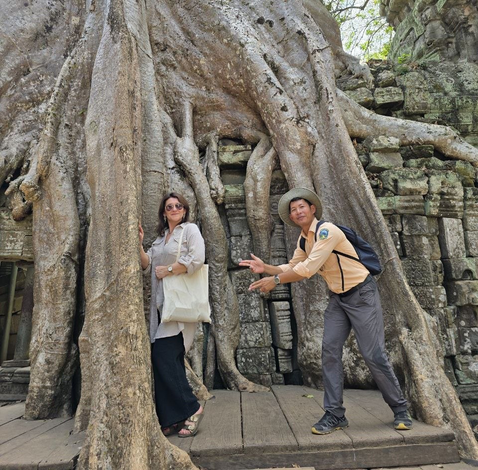 A female traveler standing next to a tour guide posing in front of massive banyan tree roots at Ta Som temple in Siem Reap