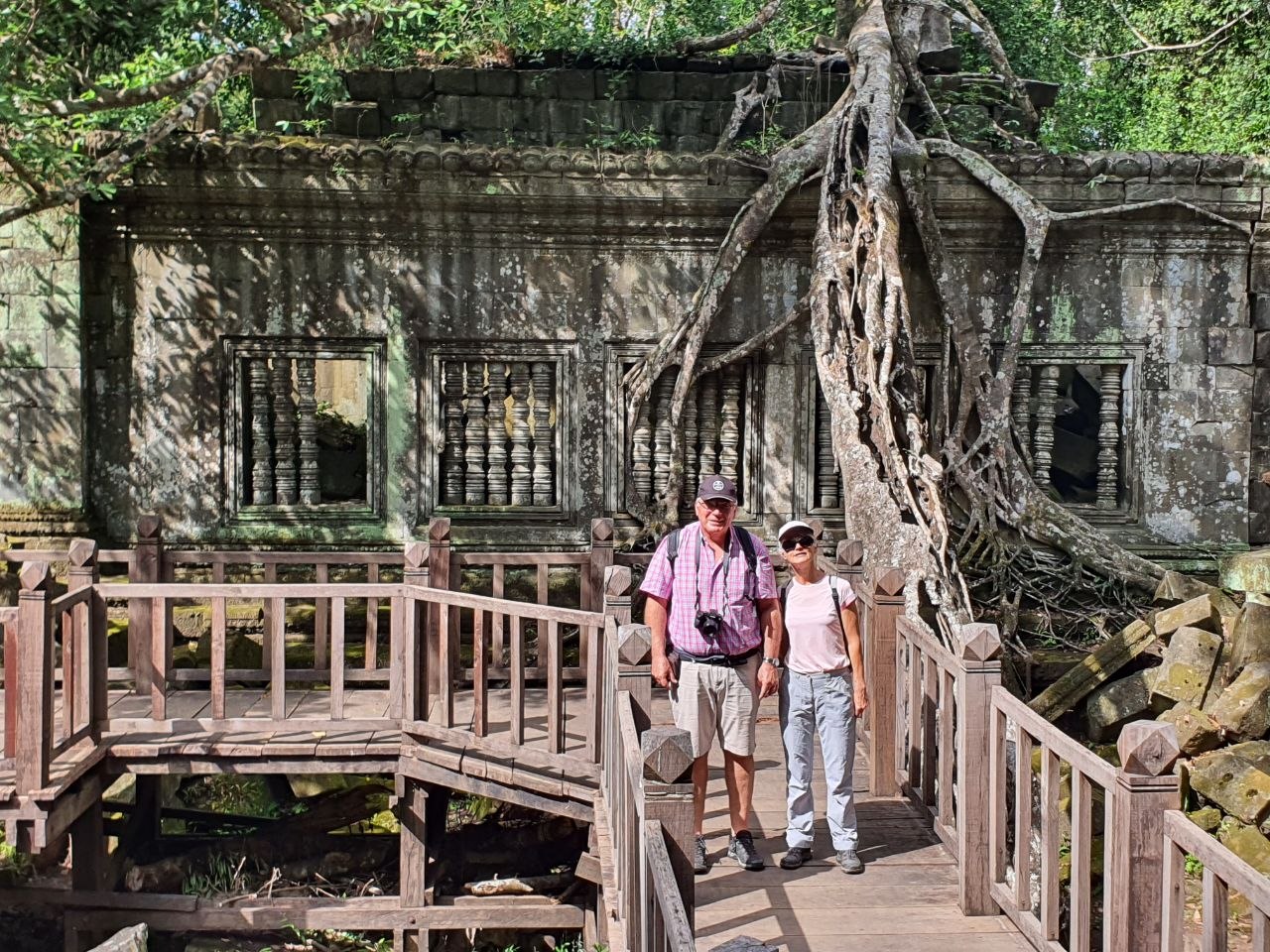 Tourists standing on a wooden boardwalk path beside overgrown temple ruins and massive tree roots at Beng Mealea