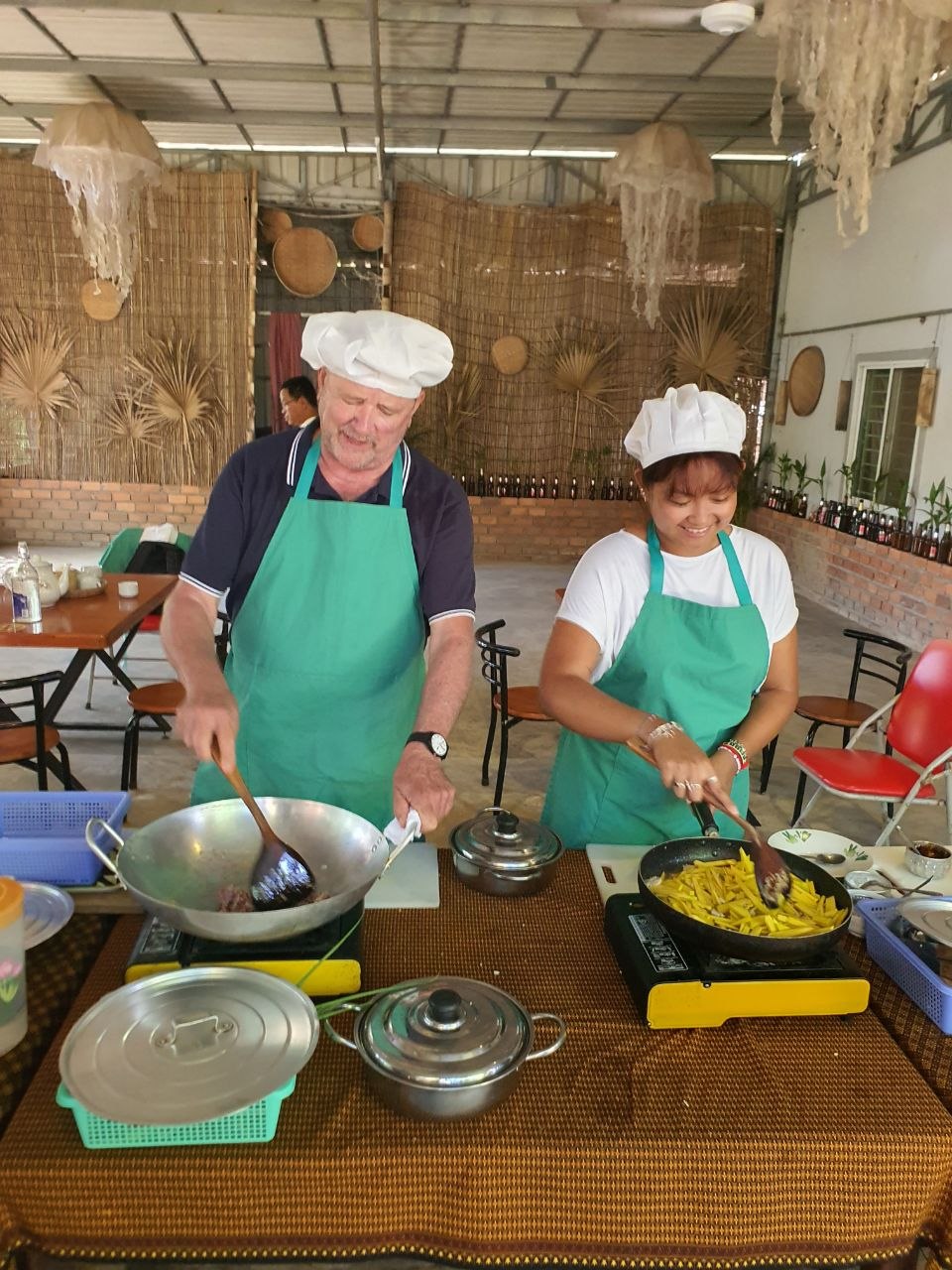 Man and woman wearing chef hats and green aprons stir-frying food during a Cambodian cooking class