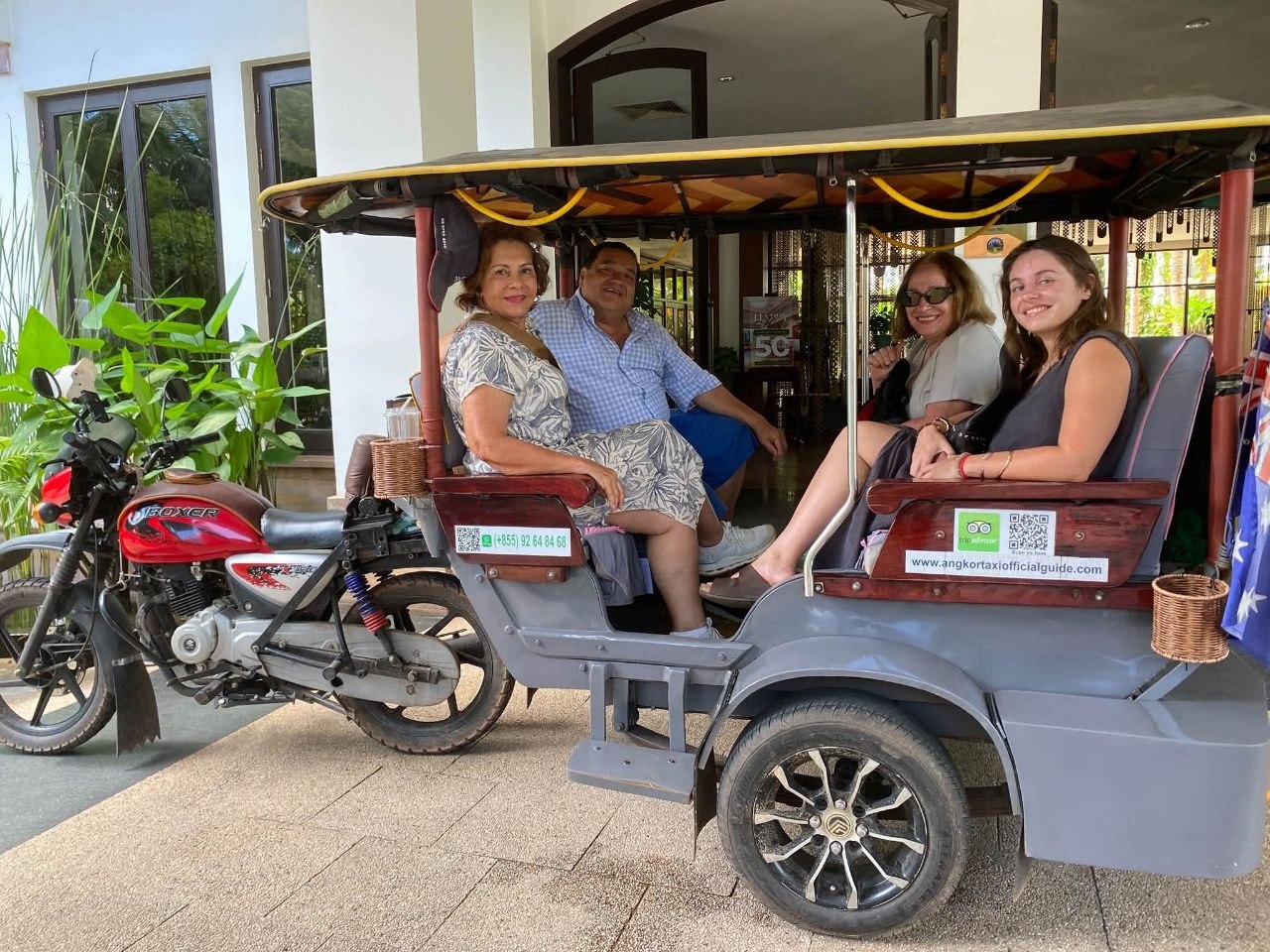 Four tourists seated inside a grey motorcycle tuk-tuk trailer outside a hotel in Siem Reap Cambodia