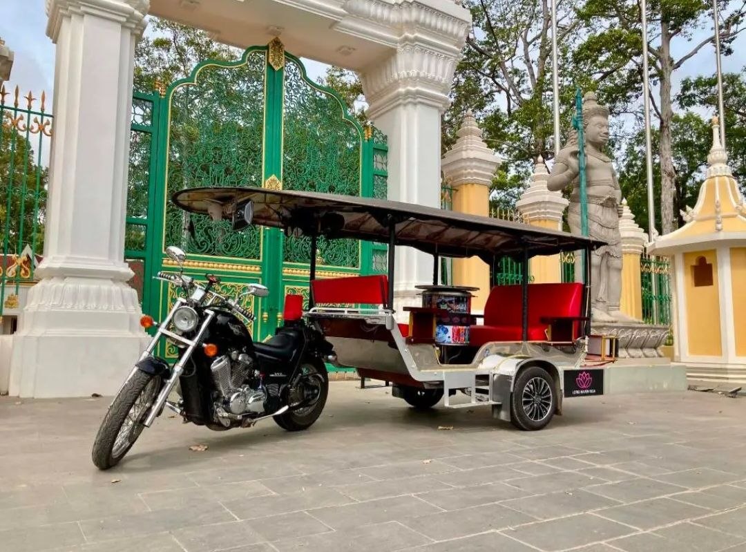 Black motorcycle with red-padded covered tuk-tuk trailer parked in front of an ornamental gate in Siem Reap Cambodia